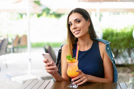 A beautiful smiling woman is drinking an exotic cocktail in an outdoor restaurant and is holding a mobile phone. Summer mood. The girl uses modern technologyの写真素材