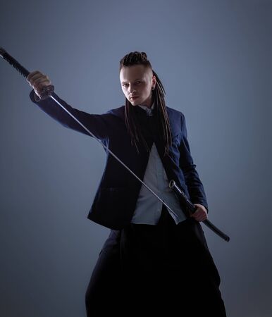 Young man dressed in the white shirt and dark blue jacket with dreadlocks holding a samurai sword. Glamour photo. Studio shooting. business man with Japanes swordの写真素材