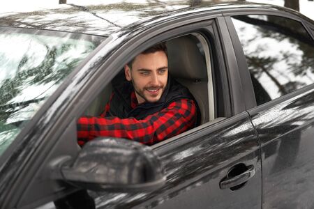 Young adult attractive Caucasian man sits at the wheel of his car sunny winter day. Wintertime road trip. Happy smiling hipster guy sitting in car and looking window. Portrait Positive driverの写真素材