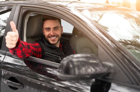 Young adult attractive Caucasian man sits at the wheel of his car sunny winter day shoeing thumbs up gesture. Wintertime road trip. Happy smiling hipster guy sitting in car and looking window.の写真素材