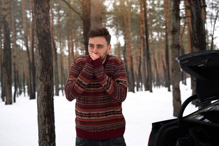 Young adult handsome man standing winter forest near his car. Attractive caucasian guy waiting for help when car is broken. Winter landscape road trip.の写真素材