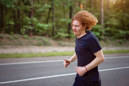 Caucasian young curly blonde man athlete runs sunny summer day on  asphalt road in the pine forest. Men 20s years jogging at the nature. training for marathon run. Healthy lifestyle concept.の写真素材