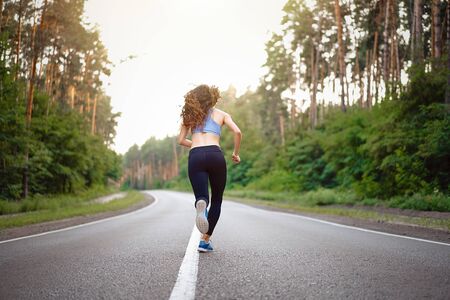 Beautiful caucasian young girl athlete runs sunny summer day on asphalt road in the pine forest view from behind. Woman 20s years jogging on nature. training for marathon. Healthy lifestyle concept.の写真素材