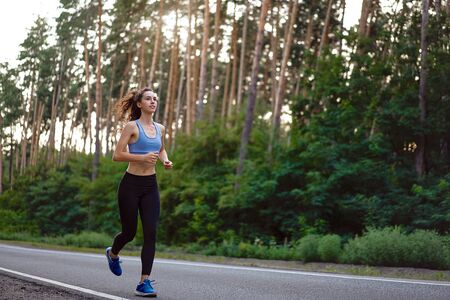 Beautiful caucasian young girl athlete runs sunny summer day on  asphalt road in the pine forest. Woman 20s years jogging at the nature. training for marathon run. Healthy lifestyle concept.の写真素材