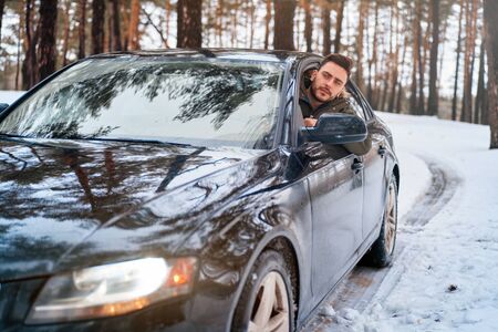 Young adult attractive Caucasian man sits at the wheel of his car sunny winter day. Wintertime road trip. Happy smiling hipster guy sitting in car and looking window. Portrait Positive driverの写真素材