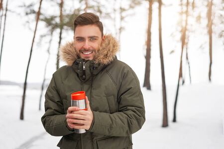 One Attractive Caucasian man stands winter forest  drink hot beverage . Winter holiday leisure. Cold season. Portrait young handsome unshaved Modern hipster guy traveling walking in winter parkの写真素材