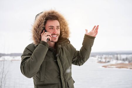 Handsome smiling brutal bearded young caucasian man standing outdoors fur hood in winter season forest Talking on a cell phone Attractive stylish european guy walking snowy christmas landscapeの写真素材