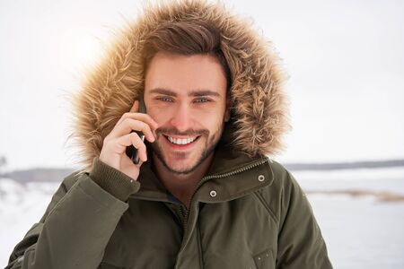 Handsome smiling brutal bearded young caucasian man standing outdoors fur hood in winter season forest Talking on a cell phone Attractive stylish european guy walking snowy christmas landscapeの写真素材
