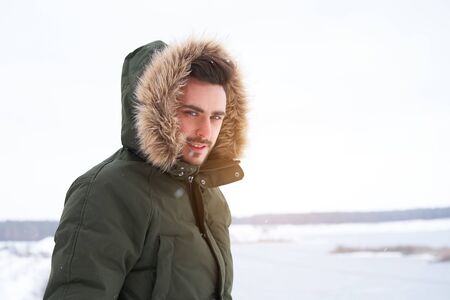 Handsome bearded young caucasian man standing outdoors fur hood winter frozen lake. Attractive stylish european guy walking snowy christmas woodland Season holiday leisureの写真素材