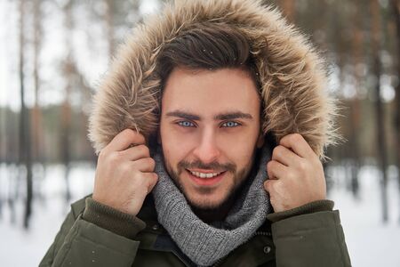 Handsome bearded young caucasian man standing outdoors fur hood in winter season forest. Attractive stylish european guy walking snowy christmas woodland Season holiday leisureの写真素材