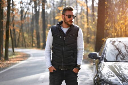 A handsome Caucasian man wearing style black vest standing near black car on nature.  Young adult...