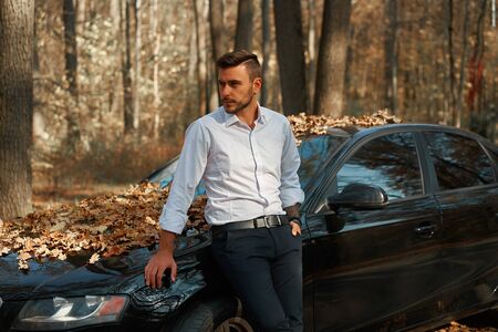 A handsome Caucasian man wearing business suit standing near black car on nature.  Young adult...