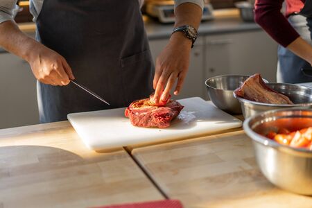 Chef in gray apron cuts meat on a plastic board. Closeup photo of the culinary process. Unrecognizable man is cooking meat in the kitchen.の写真素材