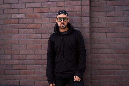 Handsome young unshaven man black hoodie and bandana stands backyard near brick walls summer day. Bearded hipster standing outdoor. Fashionable young adult caucasian guy smokes a cigarette sunglassesの写真素材