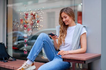 Woman city street summer sitting bench smartphone showcase shop window outdoor Modern stylish travel girl with backpack resting happy smiling Middle age female alone chatting mobile cell phoneの写真素材