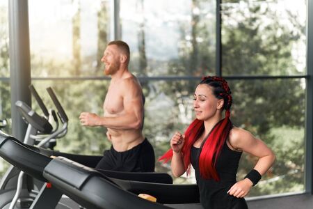 Gym treadmill running trainer man woman training together jogging fitness workout Warming up before functional cross training Personal coach train girl run on treadmil with panoramic window backgroundの写真素材