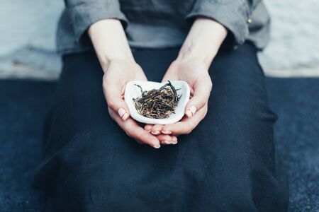 Dry tea leaves in a white saucer. Unrecognizable woman holding a bowl of tea in his hands showing it to the camera. Traditional ceremony backgroundの写真素材