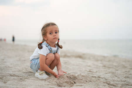 Child playing sand beach Little girl play sad alone summer family vacation Caucasian female 3 years old dressed denim near sea waterの写真素材