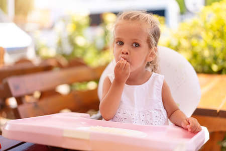 Cute little Caucasian girl eating spaghetti at table sitting in child seat outdoor restaurant. Healthy eating. Funny child eat street food sunny summer day.の写真素材