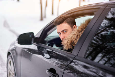Young adult attractive Caucasian man sits at the wheel of his car sunny winter day. Wintertime road...