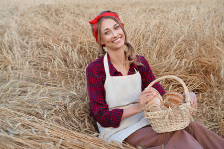Female farmer sitting wheat agricultural field Woman baker holding wicker basket bread eco product Baking small business Caucasian person dressed red plaid shirt apron organic healthy foodの写真素材