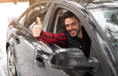 Young adult attractive Caucasian man sits at the wheel of his car sunny winter day shoeing thumbs up gesture. Wintertime road trip. Happy smiling hipster guy sitting in car and looking window.の写真素材