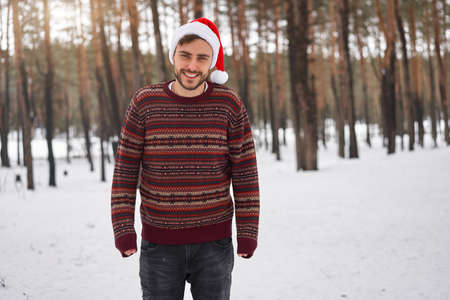 Handsome bearded young caucasian man standing outdoors sweater santa hat winter season forest. Attractive stylish european guy walking snowy christmas woodland Season holiday leisureの写真素材