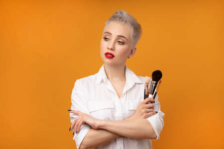 Close up portrait makeup artist. Make up courses. Concept of self visage masterclasess. Fashion professional. Woman hold makeup brushes on the hands. Studio shot on the orange color backgroundの写真素材