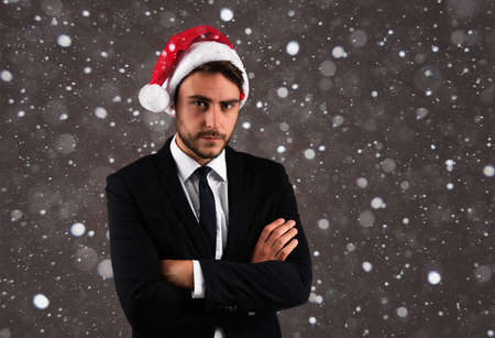 Young handsome caucasian guy in business suit and Santa hats stands on gray background in studio with serious face folded arms on chest Portrait business person with Christmas mood Holiday bannerの写真素材
