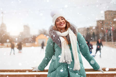 Beautiful lovely middle-aged girl curly hair warm winter jackets white knitted hat glove stands ice rink background Town Square Christmas mood lifestyle Happy holiday woman snowy day Winter leisureの写真素材