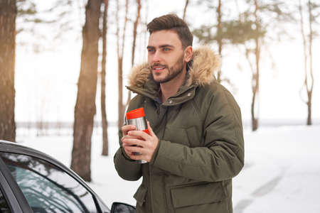 An attractive Caucasian man stands winter forest near car drink hot beverage tumbler. Winter holiday road trip. Cold season. Portrait young handsome unshaved driver. Modern hipster guy traveling on carの写真素材