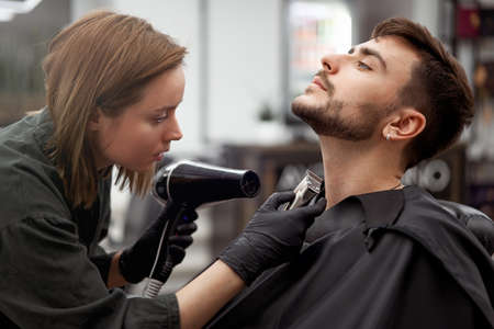 Stylish man sitting barber shop Hairstylist Hairdresser Woman blow dry his hair Portrait handsome happy young bearded caucasian guy getting trendy haircut Attractive barber girl serving clientの写真素材