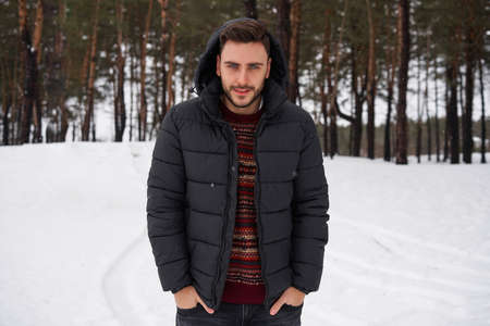 Handsome bearded young caucasian man standing outdoors in winter season forest. Attractive stylish...