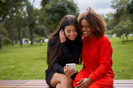Diversity and feminity concept. Happy smiling Confident young mixed race women sitting bench park outdoor Asian and african american female resting on nature summer day using smartphoneの写真素材