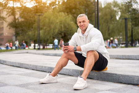 Man dressed white hoodie with coffee in hand waiting for someone in summer park Caucasian guy sitting concrete steps outdoors smilingの写真素材