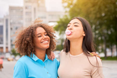African american woman laughing while asian friend inflating bubble gum. Closeup face of multiethnic friends enjoying outdoor street. Girl laughing and blowing chewing gum with friend embracing herの写真素材