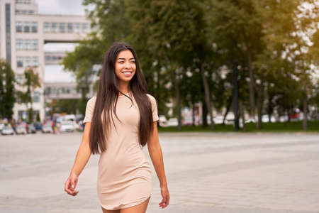 Asian girl in a beautiful tight-fitting beige dress walks through the city streets and looks away. Free space for text Fashionable female outdoors summer trendy clothesの写真素材
