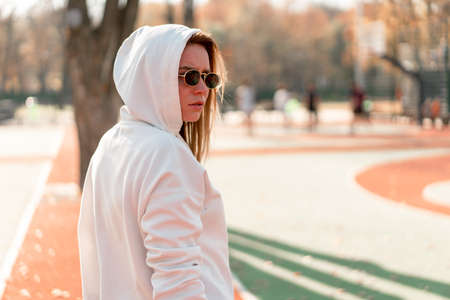 Outdoor close up portrait of young beautiful woman with in glasses, dressed in a white sweater, near the sportsground. Woman in a hood turned her head to the camera. White hoodieの写真素材