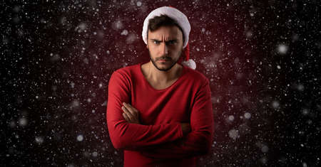 Young handsome caucasian guy in red sweater and Santa hats stands on red background in studio and feel angry. Close up portrait european guy with Christmas mood. Holiday theme. New Year.の写真素材