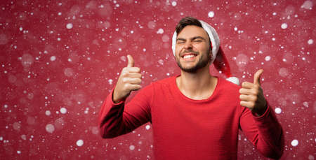 Young handsome caucasian guy in red sweater and Santa hats stands on red background in studio showing thumbs up with two hand. Close up portrait european guy with Christmas mood with snow Holiday banner.の写真素材