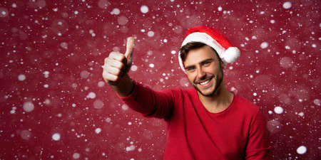 Young handsome caucasian guy in red sweater and Santa hats stands on red background in studio showing thumbs up. Close up portrait european guy with Christmas mood with snow Holiday banner.の写真素材