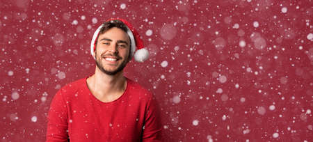 Young handsome caucasian guy in red sweater and Santa hats stands on red background in studio and teeth smiling. Close up portrait european guy with Christmas mood. Holiday banner. New Year.の写真素材