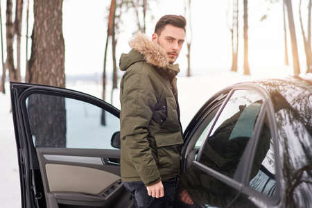 Attractive Caucasian man stands in winter forest near his car. Winter holiday road trip. Cold season. Portrait young handsome unshaved driver. Modern hipster guy traveling on carの写真素材