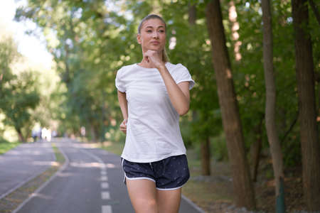 Woman running asphalt road summer park Active sporty caucasian female morning workout Healthy lifestyle concept. Athletic person sportswear exercises Dressed white t-shirtの写真素材