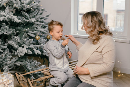 Holiday Christmas Caucasian mother playing with little cute son near decorated New year tree at home...