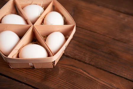 White chicken eggs lie in round wooden basket which stands on a dark wooden table. Easter...