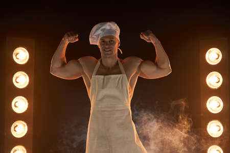 Portrait of muscular chef wearing white apron and chef hat, showing strong biceps muscle on smoky background male housewife. husband in kitchen. brutal butcher.の写真素材