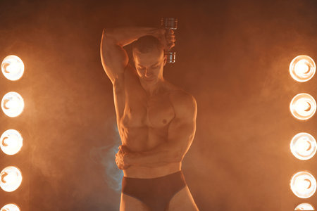 Young athlete pumping dumbbells, practicing triceps training, smoky illuminated background Healthy...