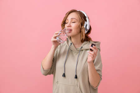 Woman with hesdphones dressed overize hoodie drink water glass bottle pink studio background Healthy lifestyle conceptの写真素材