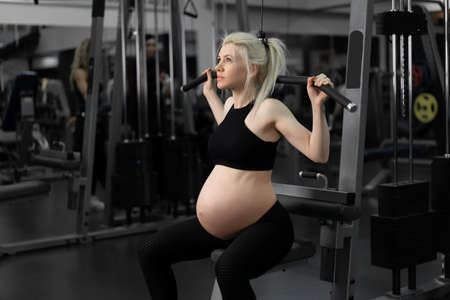 Gym workout Pregnant woman sportswear training shoulders with exercise machine Beautiful pregnancy time. Healthy life style Sport and fitness concept. Caucasian female exercising gym machineの写真素材